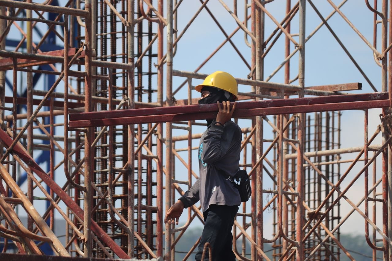 A construction worker with safety gear carrying a metal beam amidst scaffolding at a busy construction site.